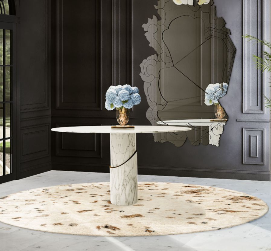 A modern round table with a white marble top and base, placed on a beige circular rug with brown accents, in a sophisticated entryway featuring dark walls and a large, ornate mirror. Two identical blue hydrangea bouquets in slender vases adorn the table.